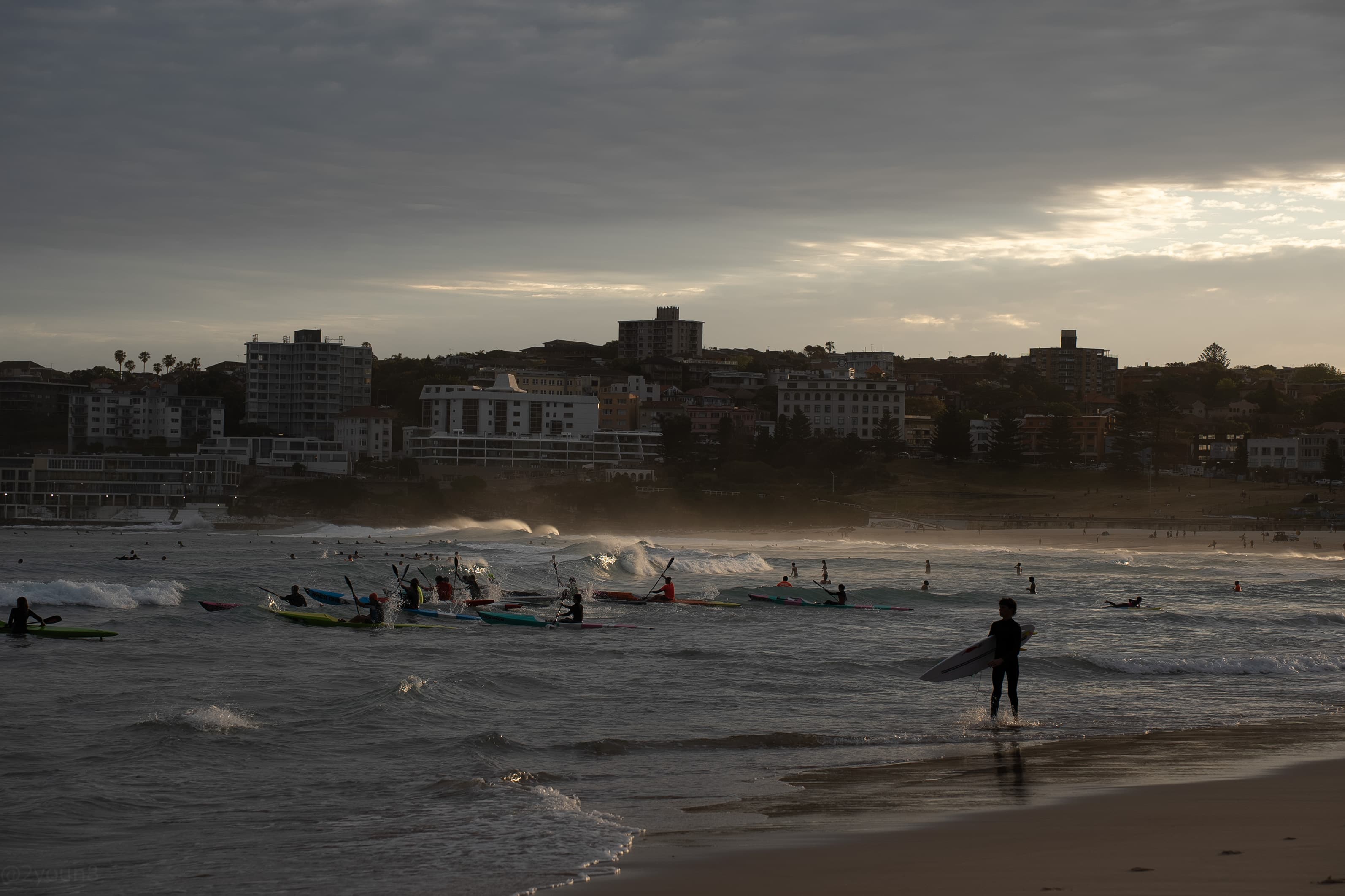A surfer waiting at the shoreline as paddlers push out behind him