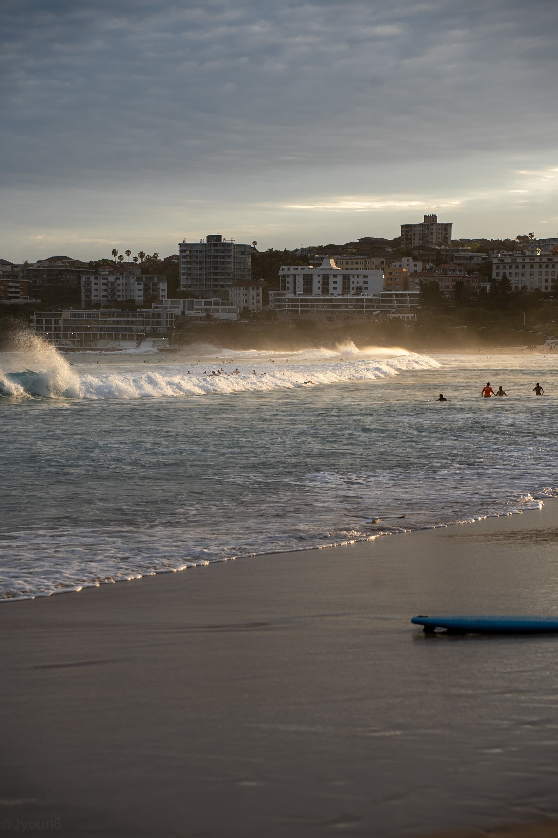 A surfboard left on the sand while swimmers wait past the break