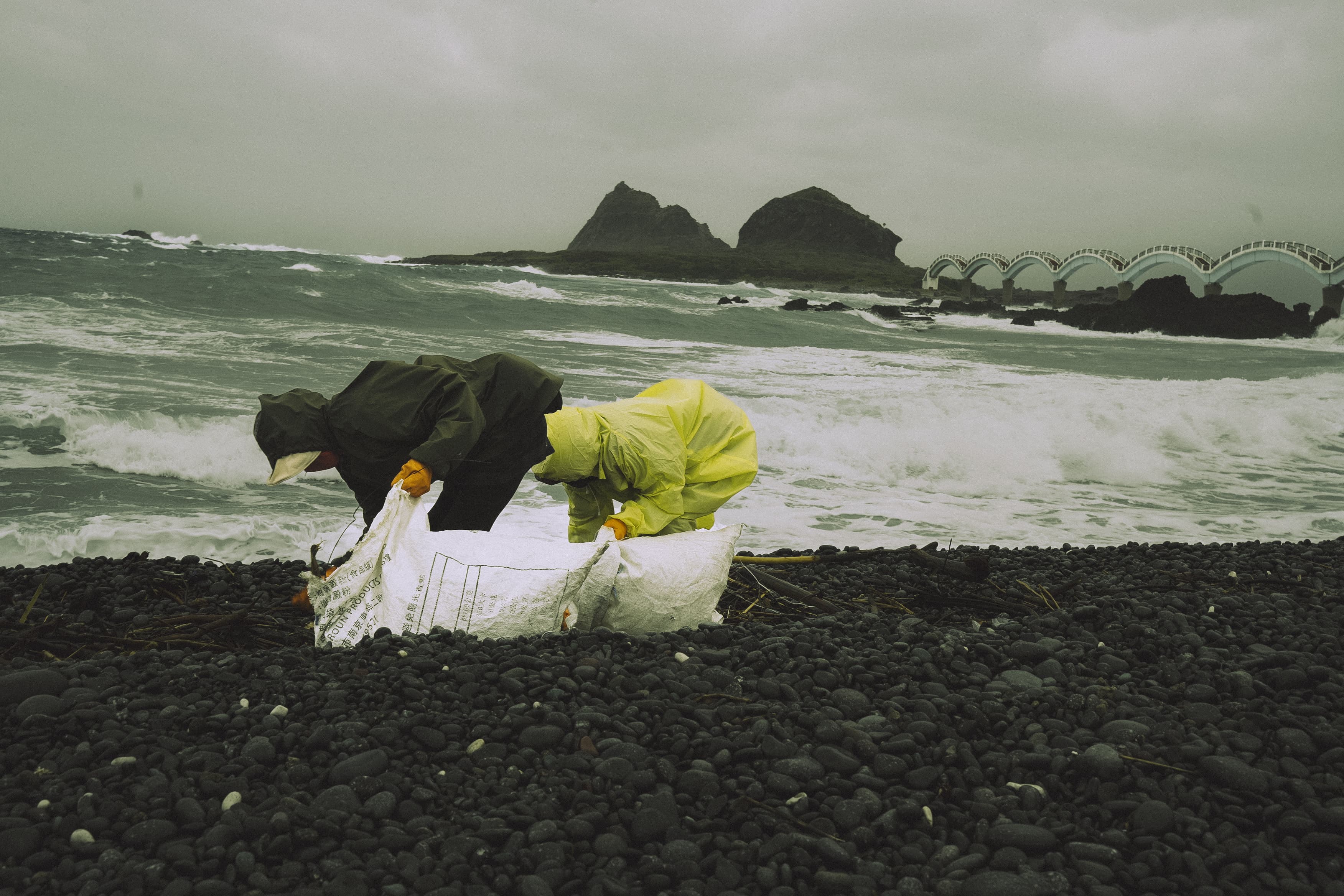 Two volunteers picking up trash on a black pebble beach